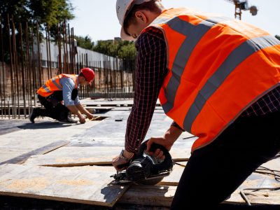 Builder dressed in orange work vest and helmet is using a circular saw on the open building site . .